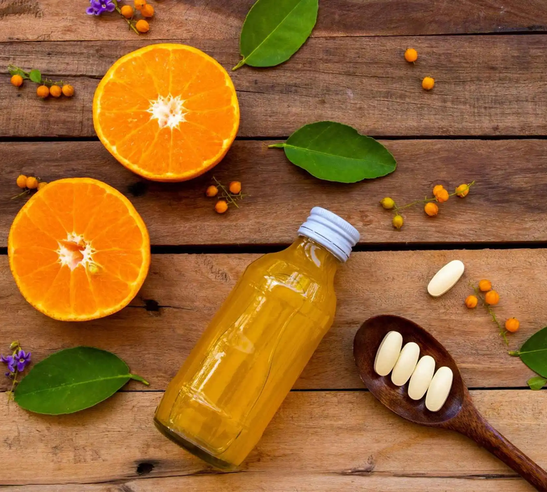 Auf einem Holztisch liegen halbierte Orangen, ein Fläschchen mit Orangensaft und ein Löffel mit Vitamin-C-Tabletten – Symbolbild für hochdosiertes Vitamin C als Nahrungsergänzung im Vergleich zur natürlichen Vitamin-C-Quelle aus Obst. | © Adobe Stock/  phenphayom