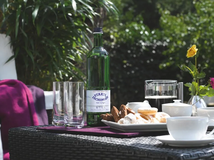 Staatl. Fachingen Still Mineralwasser in einer grünen Glasflasche auf einem gedeckten Tisch mit Kaffee, Gebäck und Gläsern im Garten. | © Fachingen Heil- und Mineralbrunnen GmbH
