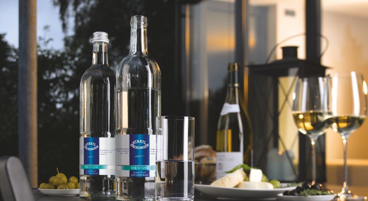 Zwei Flaschen Staatl. Fachingen Medium Mineralwasser auf einem Tisch mit Wein, Käse, Trauben und gefüllten Weingläsern bei stimmungsvoller Abendatmosphäre.