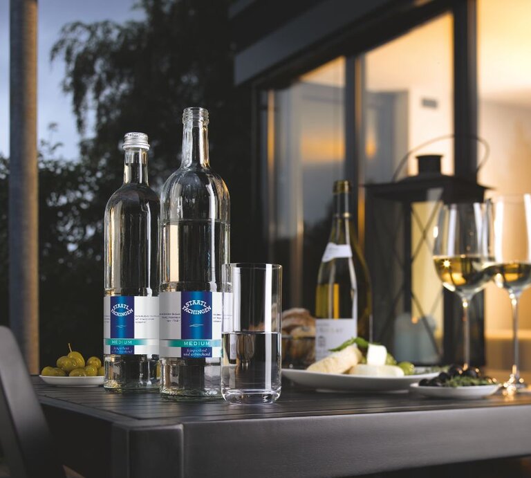 Zwei Flaschen Staatl. Fachingen Medium Mineralwasser auf einem Tisch mit Wein, Käse, Trauben und gefüllten Weingläsern bei stimmungsvoller Abendatmosphäre. | © Fachingen Heil- und Mineralbrunnen GmbH