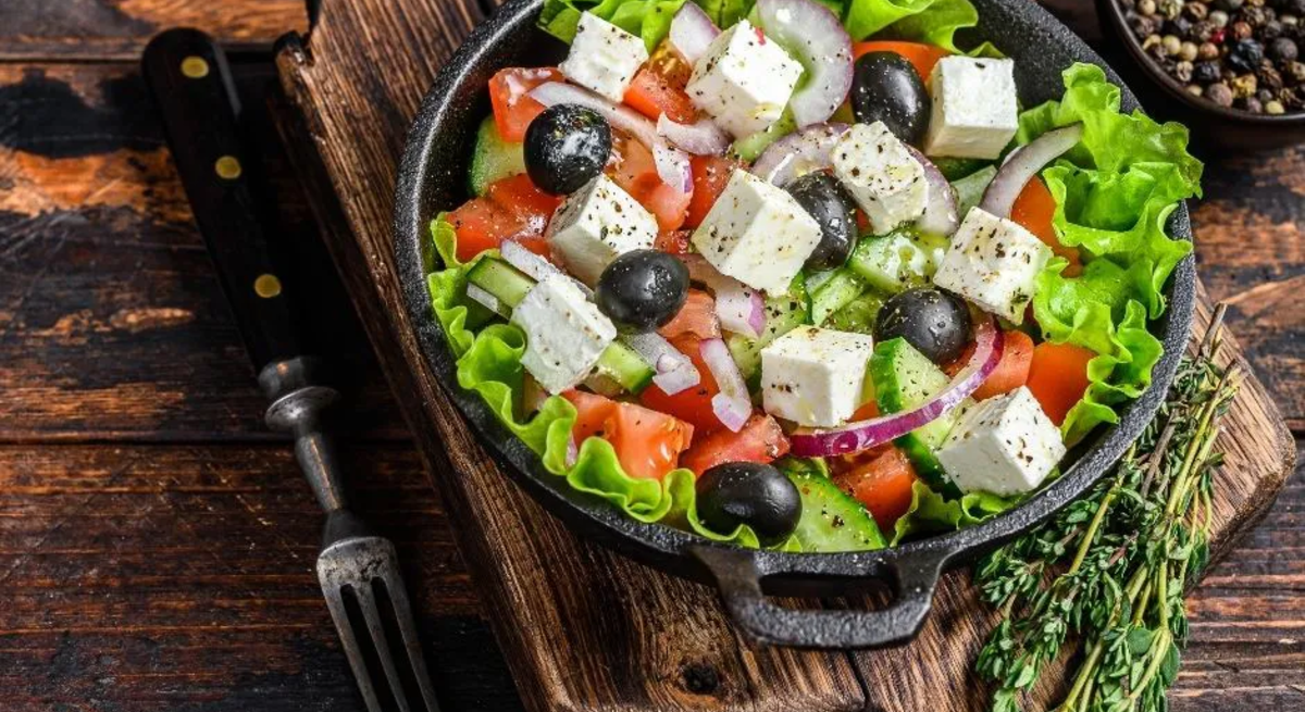 Ein frischer griechischer Salat in einer schwarzen Pfanne, serviert auf einem rustikalen Holzbrett. Der Salat besteht aus grünen Salatblättern, Tomaten, Gurkenscheiben, roten Zwiebelringen, schwarzen Oliven und gewürfeltem Feta, der leicht mit Kräutern bestreut ist. Im Hintergrund steht eine Schale mit buntem Pfeffer. Daneben liegt eine Gabel sowie ein Bund frischer Kräuter, vermutlich Thymian. Der Untergrund ist eine rustikale Holzoberfläche.