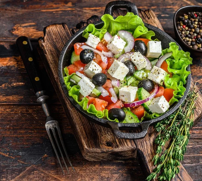 Ein frischer griechischer Salat in einer schwarzen Pfanne, serviert auf einem rustikalen Holzbrett. Der Salat besteht aus grünen Salatblättern, Tomaten, Gurkenscheiben, roten Zwiebelringen, schwarzen Oliven und gewürfeltem Feta, der leicht mit Kräutern bestreut ist. Im Hintergrund steht eine Schale mit buntem Pfeffer. Daneben liegt eine Gabel sowie ein Bund frischer Kräuter, vermutlich Thymian. Der Untergrund ist eine rustikale Holzoberfläche. | © Adobe Stock/ Vladimir Mironov