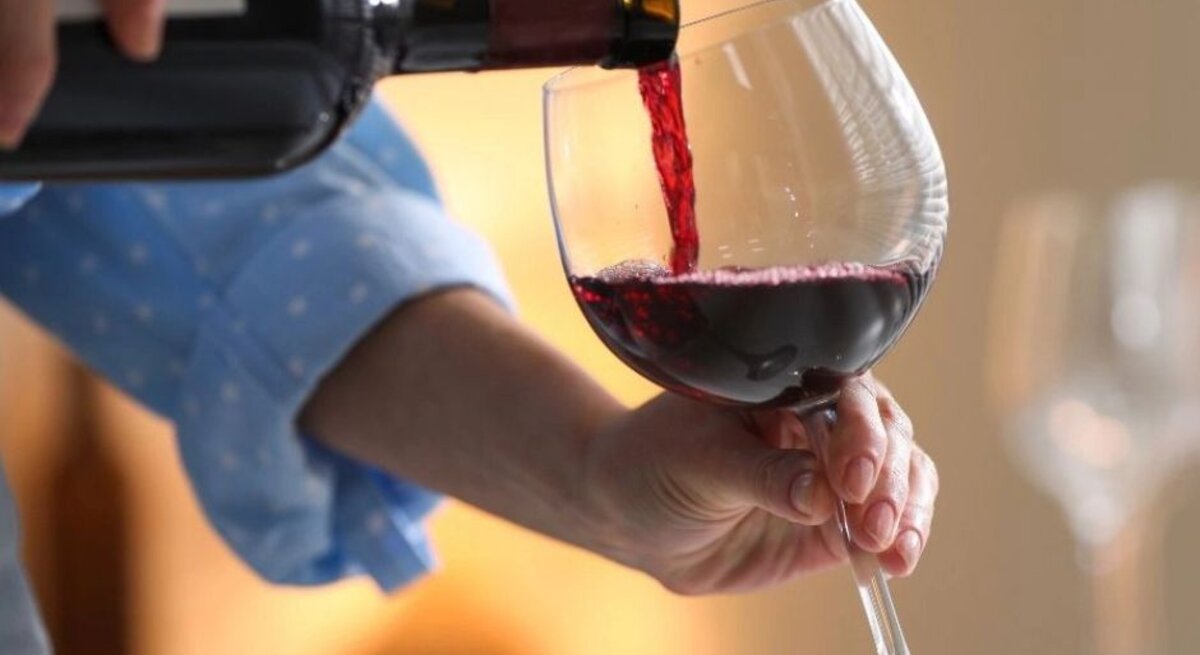 Eine Person gießt roten Wein aus einer Flasche in ein großes Weinglas; die Hand hält das Glas leicht schräg, im Hintergrund ist eine warme, wohnliche Atmosphäre zu sehen.