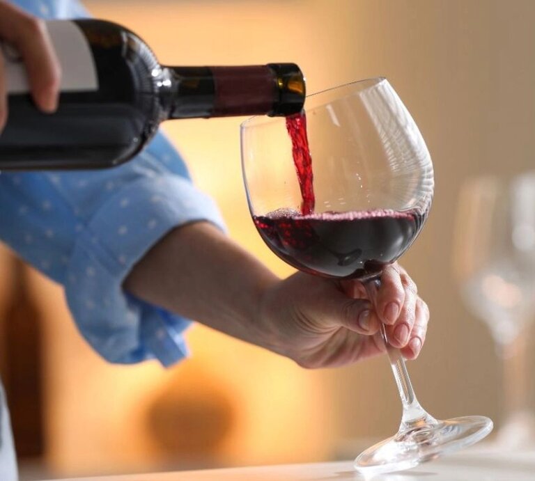 Eine Person gießt roten Wein aus einer Flasche in ein großes Weinglas; die Hand hält das Glas leicht schräg, im Hintergrund ist eine warme, wohnliche Atmosphäre zu sehen. | © Adobe Stock/ New Africa