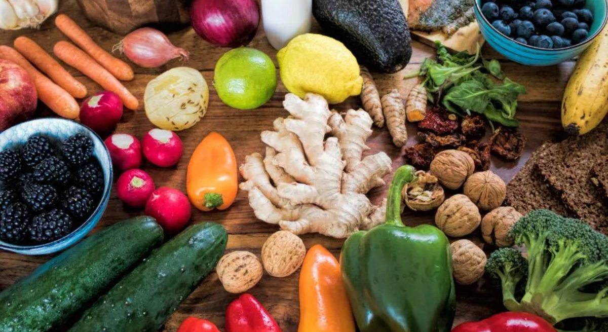 Ein Holz-Tisch mit einer Vielzahl von gesunden Lebensmitteln, darunter frisches Obst wie Zitronen, Limetten, Bananen, Blaubeeren und Brombeeren, Gemüse wie Paprika, Brokkoli, Gurken, Karotten, Rettich und Avocado, sowie weitere Zutaten wie Ingwer, Walnüsse, getrocknete Tomaten, Knoblauch, Vollkornbrot und Lachs. Im Hintergrund sind auch kleine Glasflaschen mit einer Milch- oder Joghurt-ähnlichen Flüssigkeit zu sehen.