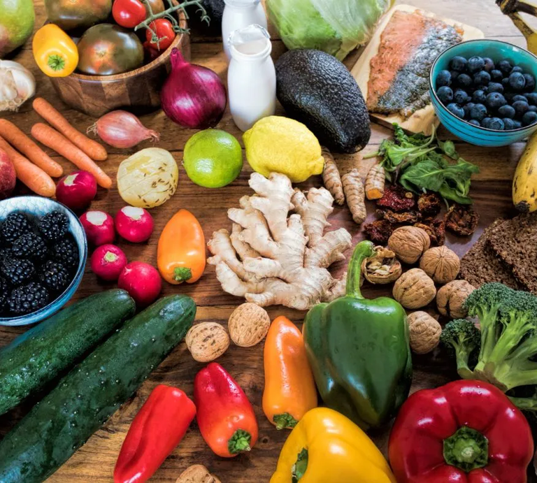 Ein Holz-Tisch mit einer Vielzahl von gesunden Lebensmitteln, darunter frisches Obst wie Zitronen, Limetten, Bananen, Blaubeeren und Brombeeren, Gemüse wie Paprika, Brokkoli, Gurken, Karotten, Rettich und Avocado, sowie weitere Zutaten wie Ingwer, Walnüsse, getrocknete Tomaten, Knoblauch, Vollkornbrot und Lachs. Im Hintergrund sind auch kleine Glasflaschen mit einer Milch- oder Joghurt-ähnlichen Flüssigkeit zu sehen. | © Adobe Stock/ ArTo