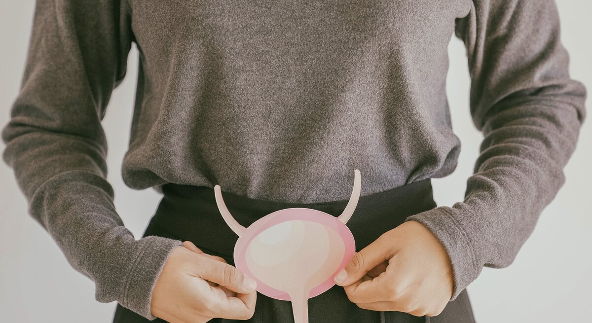 Eine Frau in einem grauen Pullover hält ein rosafarbenes, abstrahiertes Modell einer Blase vor ihrem Bauch.