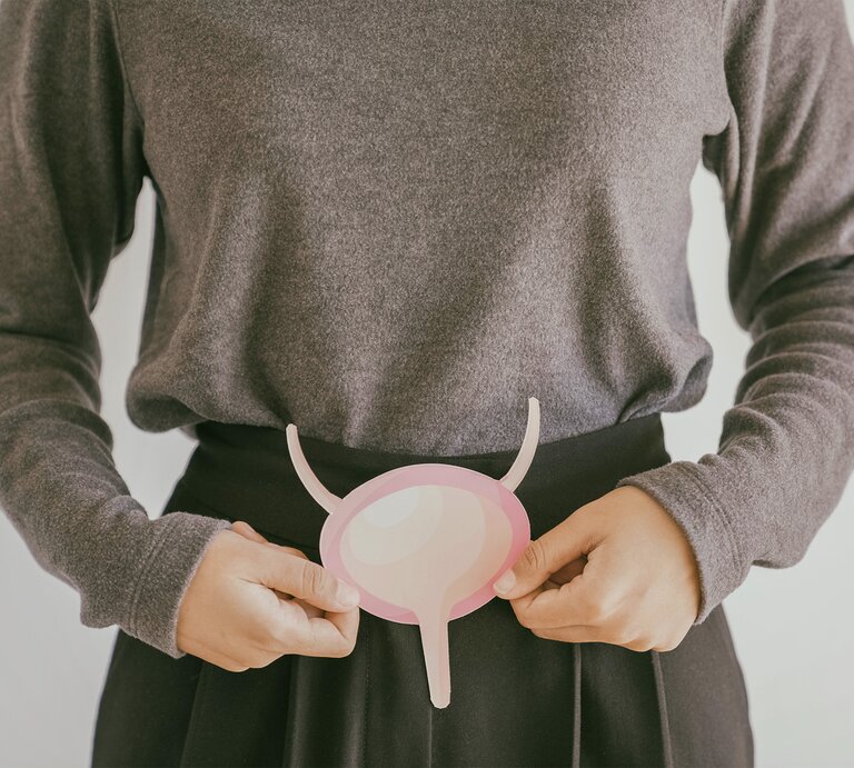 Eine Frau in einem grauen Pullover hält ein rosafarbenes, abstrahiertes Modell einer Blase vor ihrem Bauch. | © stock.adobe.com/SewcreamStudio