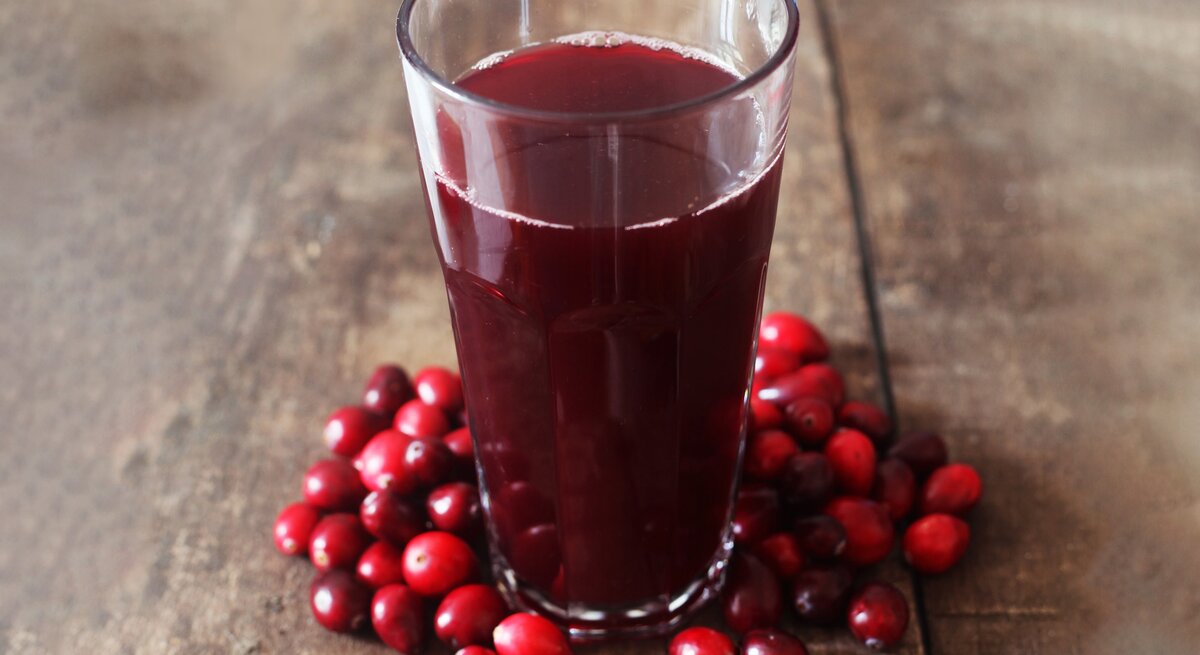 Ein Glas dunkler Cranberrysaft steht auf rustikalem Holztisch, umgeben von frischen roten Cranberries.