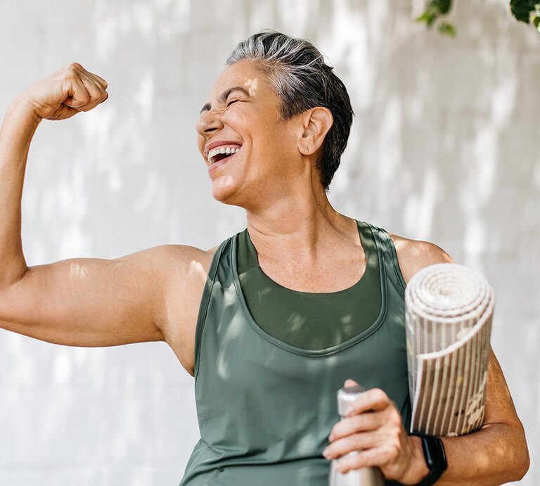 Eine lächelnde ältere Frau mit kurzen grauen Haaren zeigt stolz ihre Muskeln und hält eine Wasserflasche und Yogamatte in der Hand. | © stock.adobe.com/ (JLco) Julia Amaral