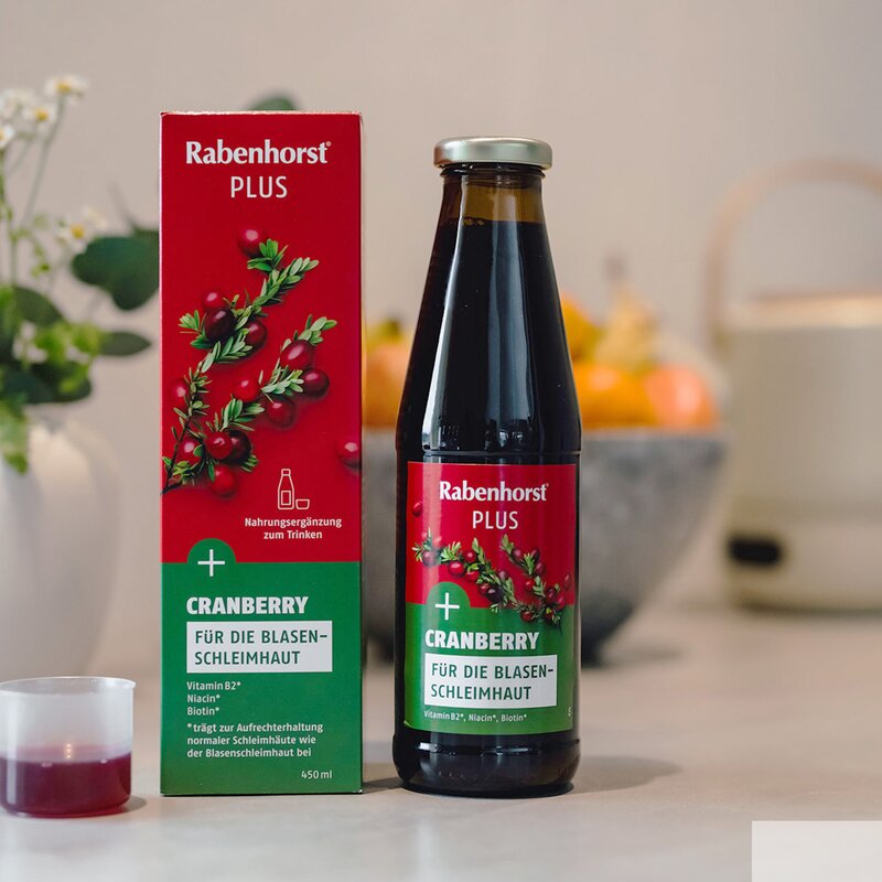 Rabenhorst Cranberry-Saft in einer Flasche und einer geöffneten Verpackung auf einem Tisch, daneben steht ein Messbecher mit rotem Saft und eine Blumenvase. | © Rabenhorst