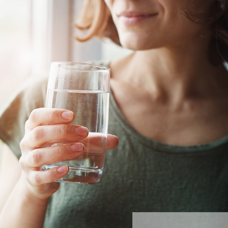 Eine junge Frau in einem grünen T-Shirt hält ein Glas mit klarem Wasser und lächelt leicht im warmen Licht des Fensters. | © stock.adobe.com/zakalinka