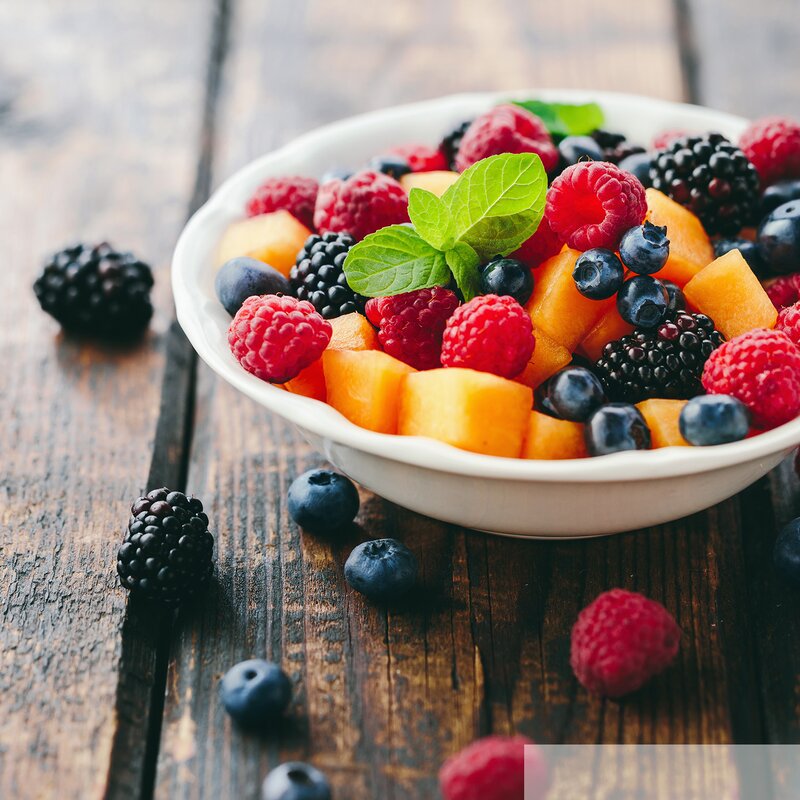 Eine weiße Schale mit frischen Beeren wie Himbeeren, Heidelbeeren und schwarzen Brombeeren, dazu Orangenstücke und ein grünes Minzblatt auf rustikalem Holztisch. | © stock.adobe.com/Melica