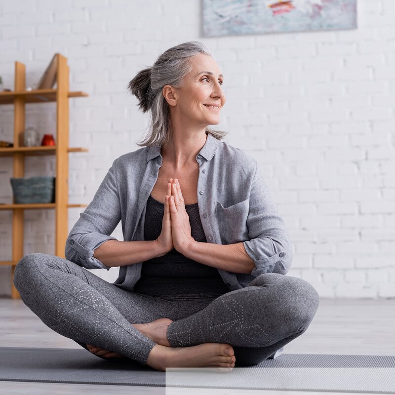 Eine Frau mit grauen Haaren trägt ein graues Shirt und sportliche Leggings, während sie in einer Yoga-Pose auf einer Matte sitzt. | © stock.adobe.com/LIGHTFIELD STUDIOS