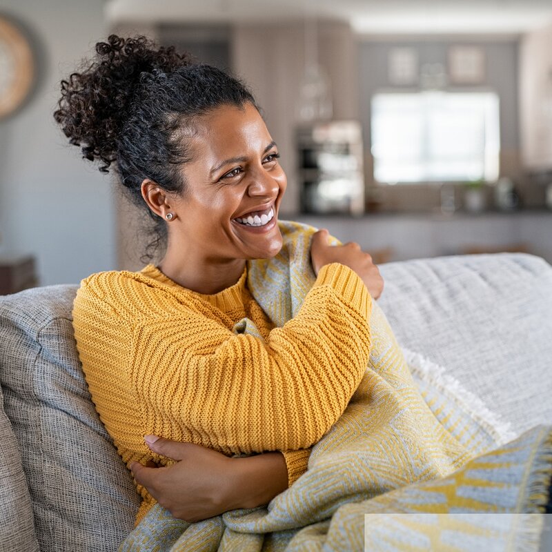 Fröhliche junge PoC Frau mit braunen, lockigen Haaren und gelbem Pullover, die sich entspannt in eine Decke gehüllt auf ihrem Sofa zurücklehnt. | © stock.adobe.com/Rido