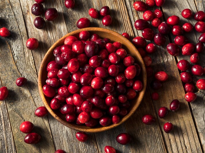  Leuchtend rote Cranberrys füllen eine runde Schale, einige rollen über die grob gemaserten Holztisch. | © stock.adobe.com/Brent Hofacker
