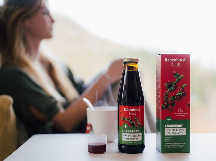 Eine Frau mit langen blonden Haaren, die in einem grünen Shirt auf einem Sessel sitzt, während auf dem Tisch ein Flasche Cranberry Plus von Rabenhorst und eine Tasse dampfend stehen. | © Rabenhorst