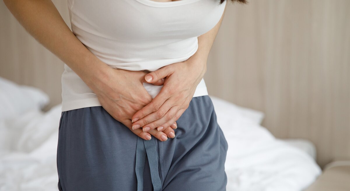 Eine junge Frau in einem weißen Top und blauen Schlafhosen hält sich schmerzerfüllt den Bauch, während sie vor dem Bett steht.