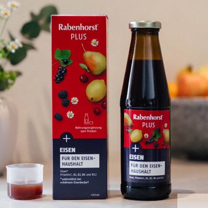 Rabenhorst PLUS Eisen in Glasflasche und roter Verpackung auf hellem Tisch mit Dekovase und Schale voller Äpfel im Hintergrund. | © Rabenhorst