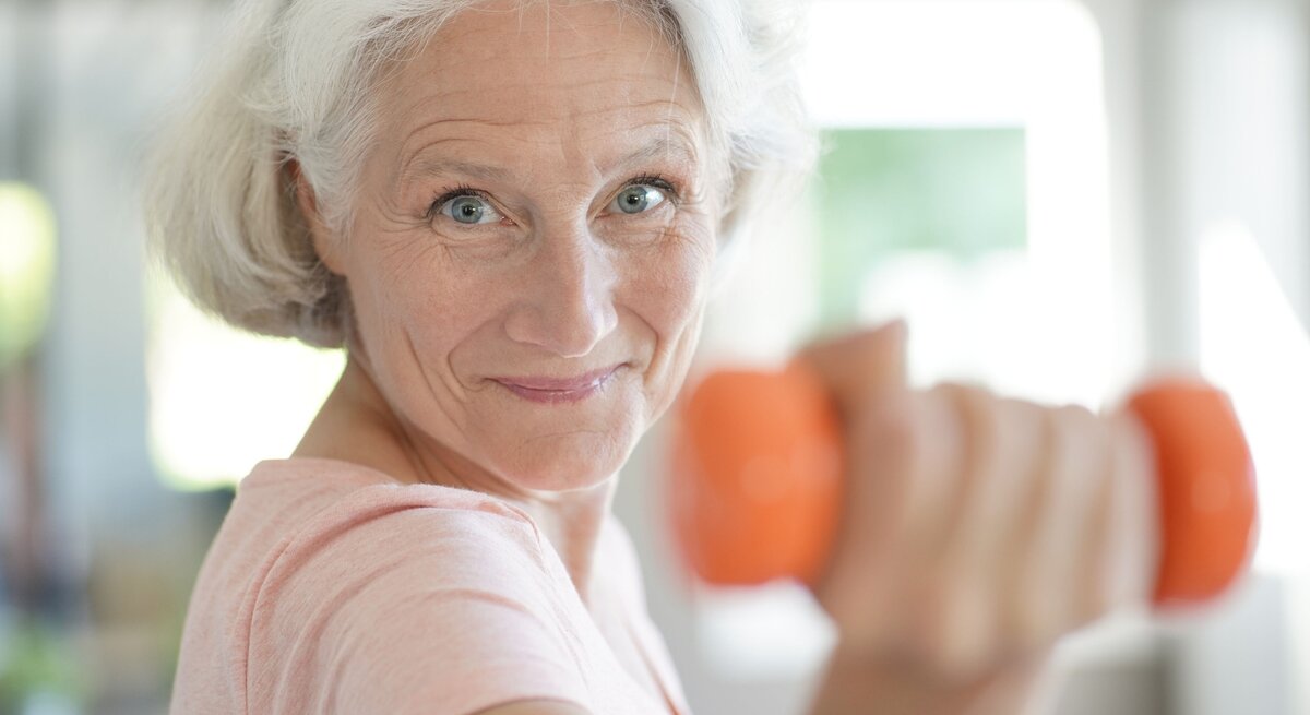 Ältere Frau mit grauem Haar und rosa T-Shirt hält eine orangefarbene Hantel und lächelt motiviert in die Kamera. Sie scheint sich in einem hellen, modernen Raum zu befinden und wirkt aktiv und gesund.