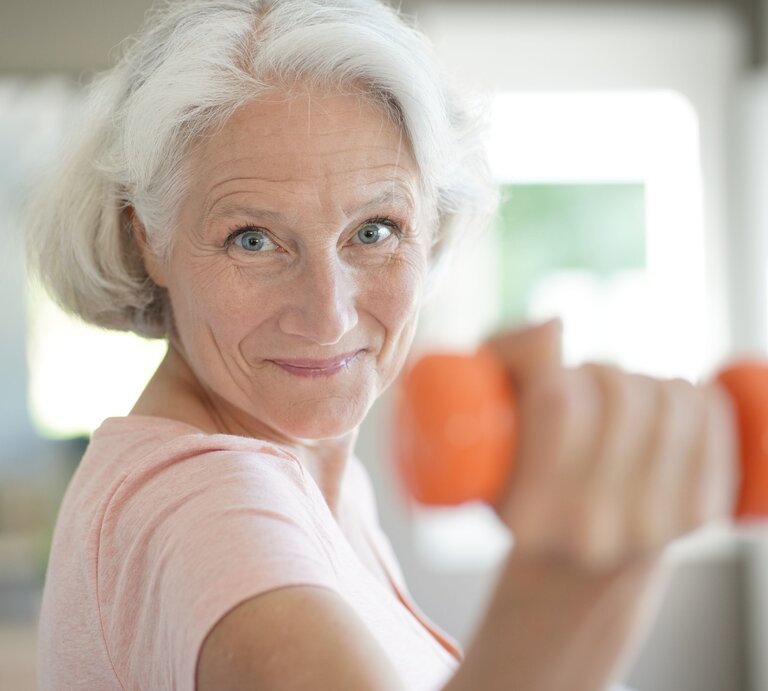 Ältere Frau mit grauem Haar und rosa T-Shirt hält eine orangefarbene Hantel und lächelt motiviert in die Kamera. Sie scheint sich in einem hellen, modernen Raum zu befinden und wirkt aktiv und gesund. | © Adobe Stock/ goodluz