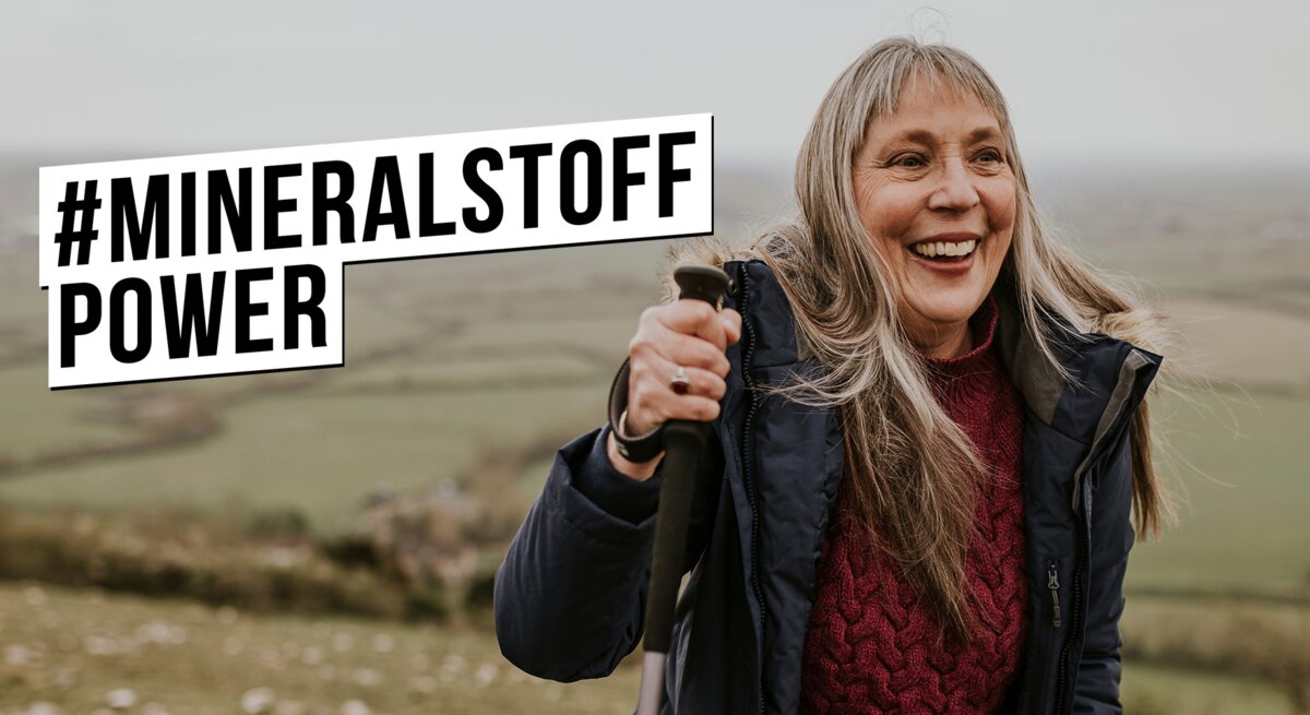 Lachende ältere Frau mit langen grauen Haaren wandert mit Stöcken im Wollpullover und Winterjacke über grünen Hügel.