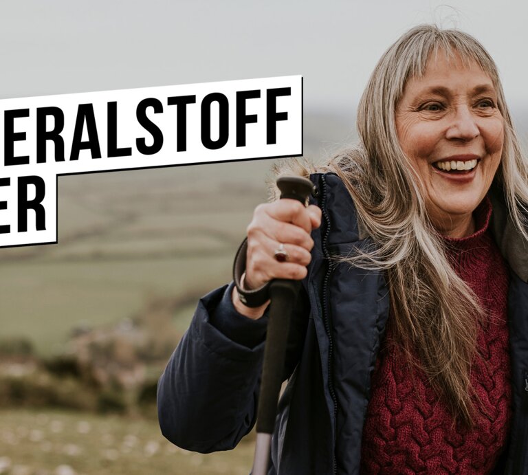 Lachende ältere Frau mit langen grauen Haaren wandert mit Stöcken im Wollpullover und Winterjacke über grünen Hügel. | © Rawpixel.com - stock.adobe.com