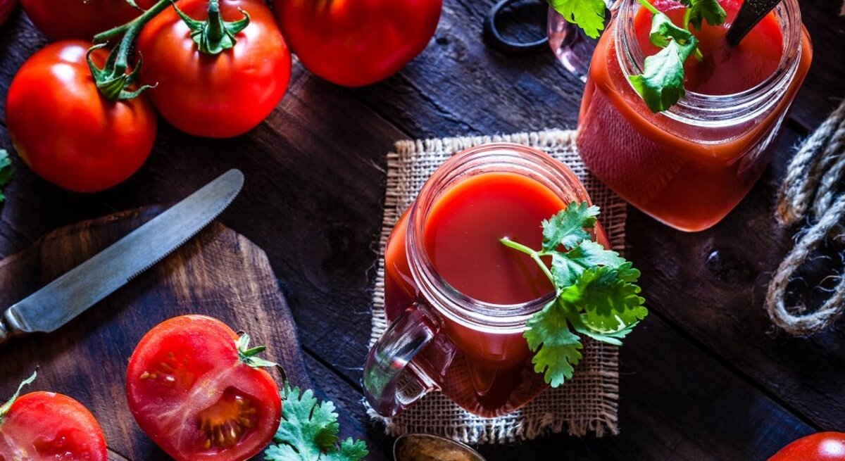 Ein Tisch aus Holz mit einem Brettchen, auf dem ein Messer liegt. Tomaten liegen auf dem Tisch und ein Glas mit Tomatensaft ist von Oben zu sehen. 