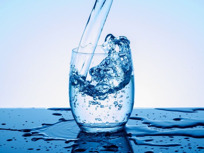 Klares Wasser wird in ein transparentes Glas gegossen, dabei spritzt es dynamisch über den Rand; das Glas steht auf einer nassen Oberfläche vor einem hellblauen Hintergrund. | © Getty Images/ K-Paul via Canva.com