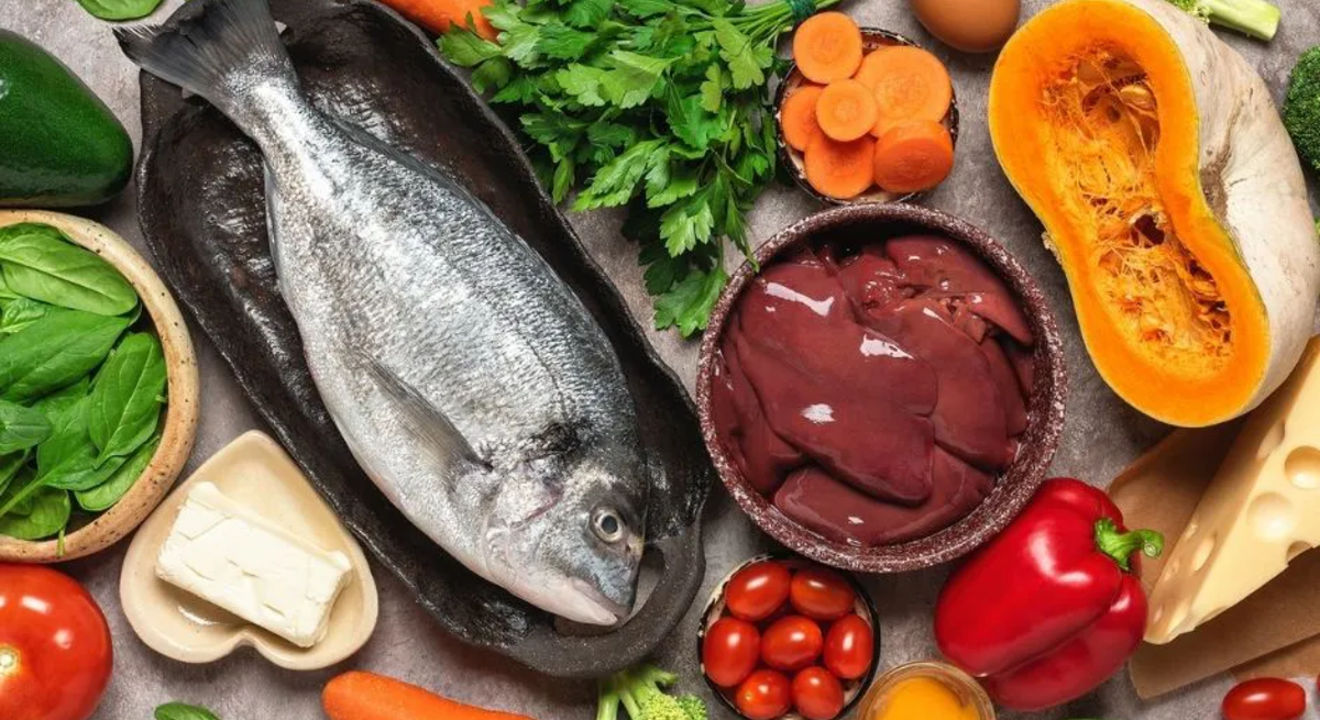 Ein Stillleben mit einer Auswahl an Lebensmitteln, die reich an Nährstoffen sind. Zu sehen sind ein ganzer Fisch auf einem dunklen Teller, eine Schale mit Leberstücken, frisches Gemüse wie Karotten, Paprika, Brokkoli, Spinat, eine Avocado und eine halbierte Kürbishälfte. Außerdem gibt es kleine Tomaten, frische Petersilie, ein Ei, ein Stück Butter und ein Stück Käse. Alles liegt auf einem grauen Untergrund arrangiert.