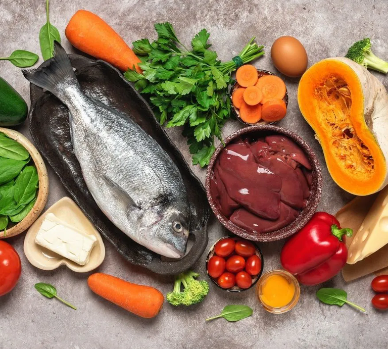 Ein Stillleben mit einer Auswahl an Lebensmitteln, die reich an Nährstoffen sind. Zu sehen sind ein ganzer Fisch auf einem dunklen Teller, eine Schale mit Leberstücken, frisches Gemüse wie Karotten, Paprika, Brokkoli, Spinat, eine Avocado und eine halbierte Kürbishälfte. Außerdem gibt es kleine Tomaten, frische Petersilie, ein Ei, ein Stück Butter und ein Stück Käse. Alles liegt auf einem grauen Untergrund arrangiert. | © GettyImages/Yulia Gusterina