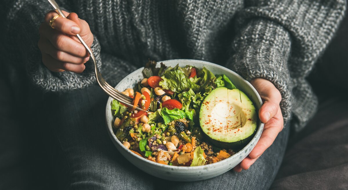 Das Bild zeigt eine Nahaufnahme einer Person, die eine Schale mit einem bunten und gesunden veganen Salat hält. Die Person trägt einen gemütlichen grauen Strickpullover und ist in eine entspannte Haltung versunken, während sie mit einer Gabel in der rechten Hand zum Essen ansetzt. Der Salat besteht aus einer Vielzahl von Zutaten, darunter grüne Blätter, Kichererbsen, Tomaten, Avocado und Körner, die alle frisch und ansprechend aussehen. Die Avocado im Vordergrund ist reif und aufgeschnitten, mit einem Spritzer Gewürz oben drauf. Der Fokus auf den Salat und die Handlungen der Person spiegelt die Wichtigkeit einer achtsamen und gesundheitsbewussten Ernährung wider.