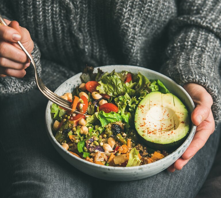Das Bild zeigt eine Nahaufnahme einer Person, die eine Schale mit einem bunten und gesunden veganen Salat hält. Die Person trägt einen gemütlichen grauen Strickpullover und ist in eine entspannte Haltung versunken, während sie mit einer Gabel in der rechten Hand zum Essen ansetzt. Der Salat besteht aus einer Vielzahl von Zutaten, darunter grüne Blätter, Kichererbsen, Tomaten, Avocado und Körner, die alle frisch und ansprechend aussehen. Die Avocado im Vordergrund ist reif und aufgeschnitten, mit einem Spritzer Gewürz oben drauf. Der Fokus auf den Salat und die Handlungen der Person spiegelt die Wichtigkeit einer achtsamen und gesundheitsbewussten Ernährung wider. | © Adobe Stock/ sonyakamoz