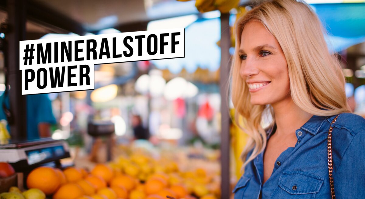 Junge blonde Frau in Jeanshemd lächelt zufrieden beim Auswählen von Birnen an einem bunten Obststand auf dem Wochenmarkt.