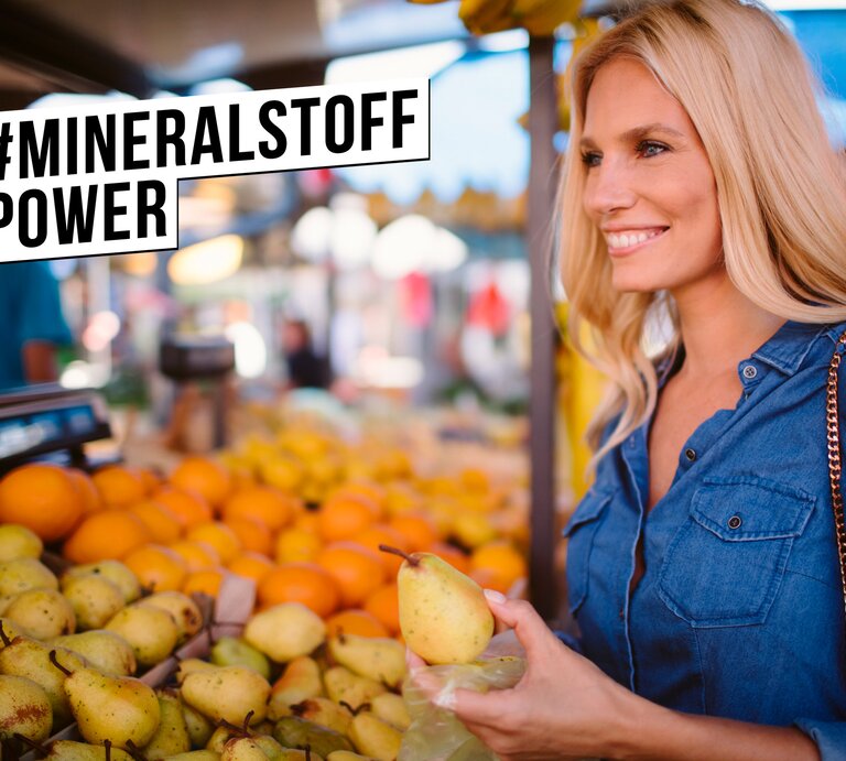 Junge blonde Frau in Jeanshemd lächelt zufrieden beim Auswählen von Birnen an einem bunten Obststand auf dem Wochenmarkt. | © istock.com/vgajic