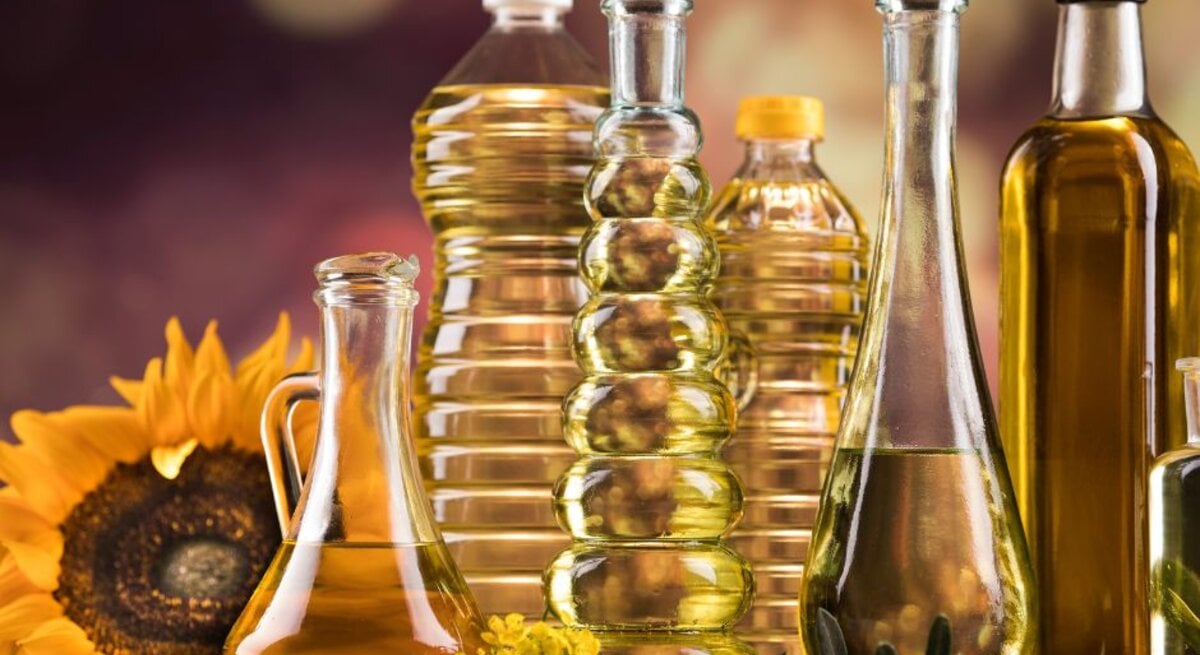 Verschiedene Flaschen mit Speiseöl aus Glas und Kunststoff stehen nebeneinander auf einem Tisch, darunter Sonnenblumenöl und Olivenöl; im Hintergrund sind eine Sonnenblume und warme, unscharfe Farben zu sehen.