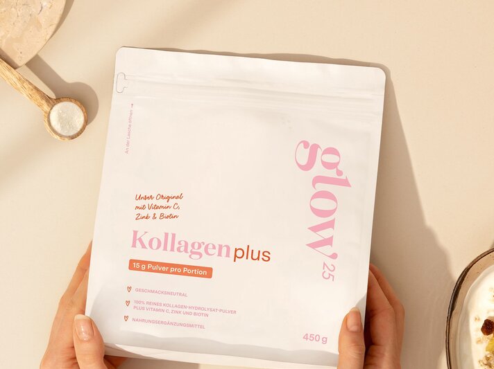 Eine helle Verpackung Kollagenpulver wird von zwei Händen gehalten, daneben liegen Holzlöffel mit Salz und eine Schale Müsli auf beigem Tisch. | © Glow25