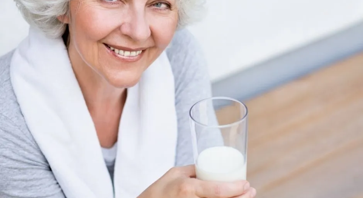 Ältere Frau mit grauen Haaren lächelt und hält ein Glas Milch in der Hand – natürliche Quelle für Vitamin B12.