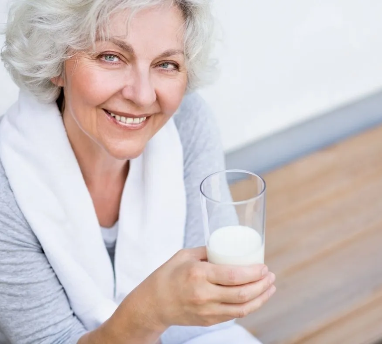 Ältere Frau mit grauen Haaren lächelt und hält ein Glas Milch in der Hand – natürliche Quelle für Vitamin B12. | © Adobe Stock/ jd-photodesign