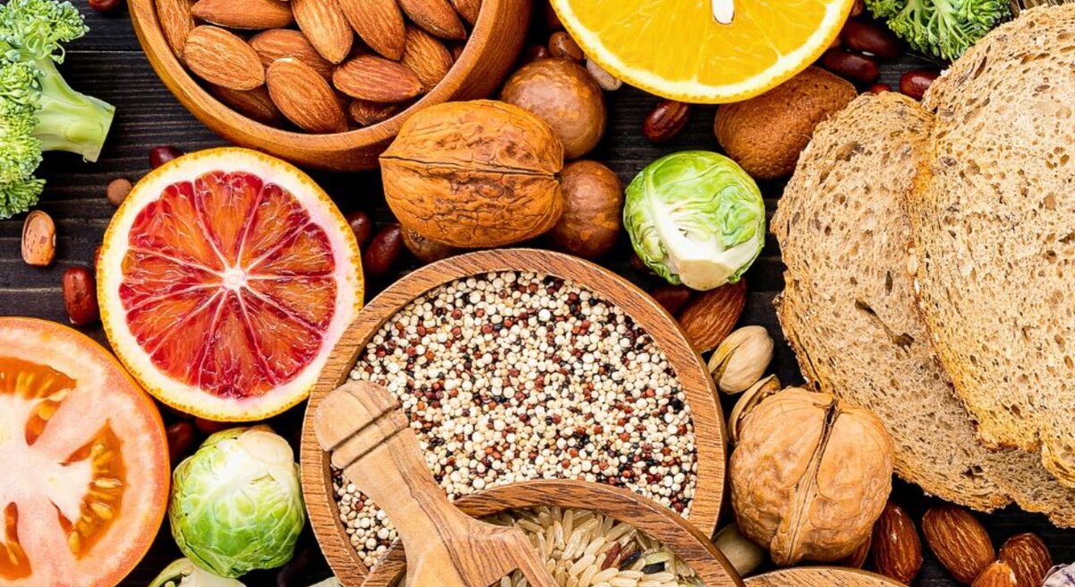 Verschiedene ballaststoffreiche Lebensmittel wie Vollkornbrot, Nüsse, Samen, Reis, Quinoa, Hülsenfrüchte, Brokkoli, Rosenkohl, Karotten und Zitrusfrüchte liegen dekorativ auf einem dunklen Untergrund.