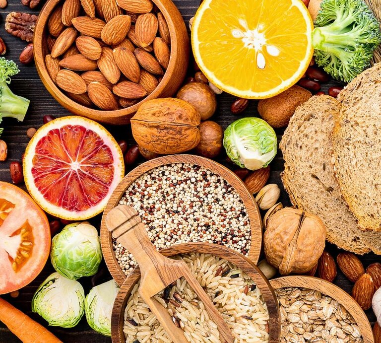 Verschiedene ballaststoffreiche Lebensmittel wie Vollkornbrot, Nüsse, Samen, Reis, Quinoa, Hülsenfrüchte, Brokkoli, Rosenkohl, Karotten und Zitrusfrüchte liegen dekorativ auf einem dunklen Untergrund. | © Diaconescu Vlad's Images via Canva.com