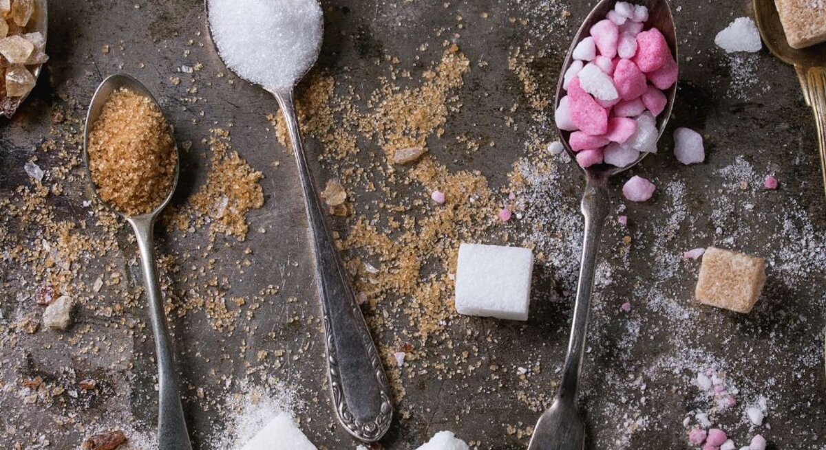 Mehrere Löffel mit unterschiedlicher Zuckerart – weißer Zucker, brauner Zucker und pink-weiße Zuckerstückchen – liegen auf einer dunklen Oberfläche, umgeben von verstreuten Zuckerwürfeln und -krümeln.