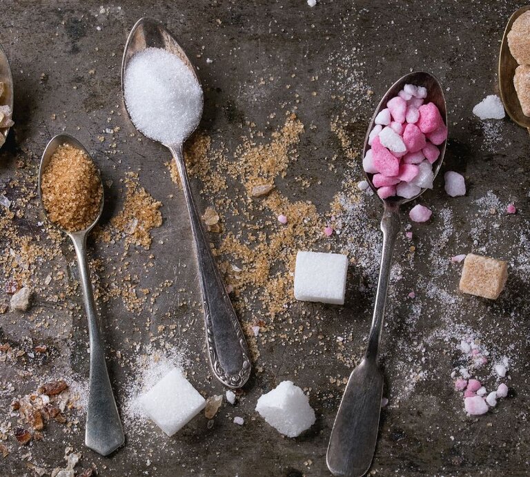 Mehrere Löffel mit unterschiedlicher Zuckerart – weißer Zucker, brauner Zucker und pink-weiße Zuckerstückchen – liegen auf einer dunklen Oberfläche, umgeben von verstreuten Zuckerwürfeln und -krümeln. | © natashabreen via Canva. com 