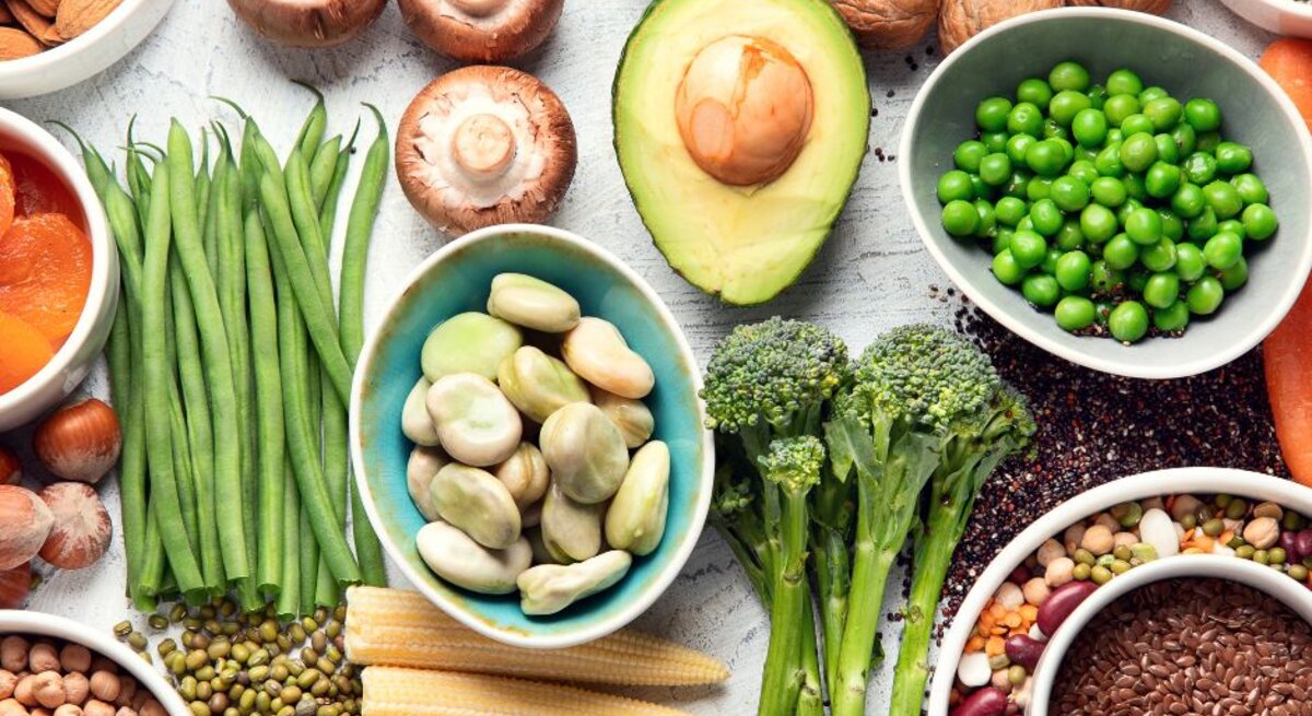 Von oben fotografierte Vielfalt pflanzlicher Lebensmittel—Erbsen, Bohnen, Linsen, Avocado, Brokkoli, Pilze, grüne Bohnen, Nüsse, Samen und Babymais—auf hellem Untergrund.