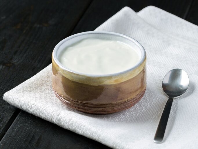Eine kleine, rustikale Keramikschale mit Skyr auf einer weißen Stoffserviette; daneben liegt ein Teelöffel auf dunkler Holzoberfläche. | © Getty Images/ Catto32 via Canva.com