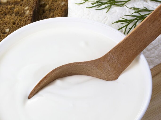 Nahaufnahme einer weißen Schüssel mit glattem, cremigem Quark, in den ein hölzerner Löffel eintaucht; im Hintergrund dunkles Brot, frisches Dillgrün und ein weißer Frischkäse-Laib. | © magnola via Canva.com