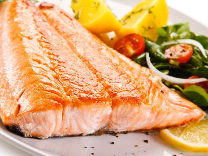 Gebratenes Lachsfilet mit glänzender, leicht karamellisierter Oberfläche auf einem Teller, serviert mit Zitronenscheiben, Kartoffelspalten und grünem Salat mit Tomaten und Zwiebeln. | © Getty Images Pro/ gbh007 via Canva.com