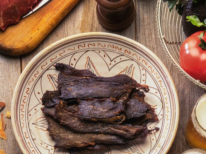 Teller mit dunkelbraunen, getrockneten Fleischstreifen (Beef Jerky) in rustikalem Setting; daneben Gewürze, frisches Gemüse und ein Glas Getränk auf einem Holztisch. | © Aflo Images via Canva.com