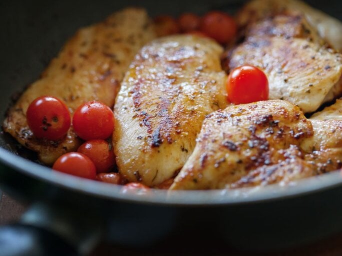 Gebratene Hähnchenbrustfilets mit goldbrauner, gewürzter Kruste in einer Pfanne, dazwischen ganze Kirschtomaten; Nahaufnahme mit sichtbar glänzenden Röstaromen. | © Getty Images Signature/ anandaBGD via Canva.com