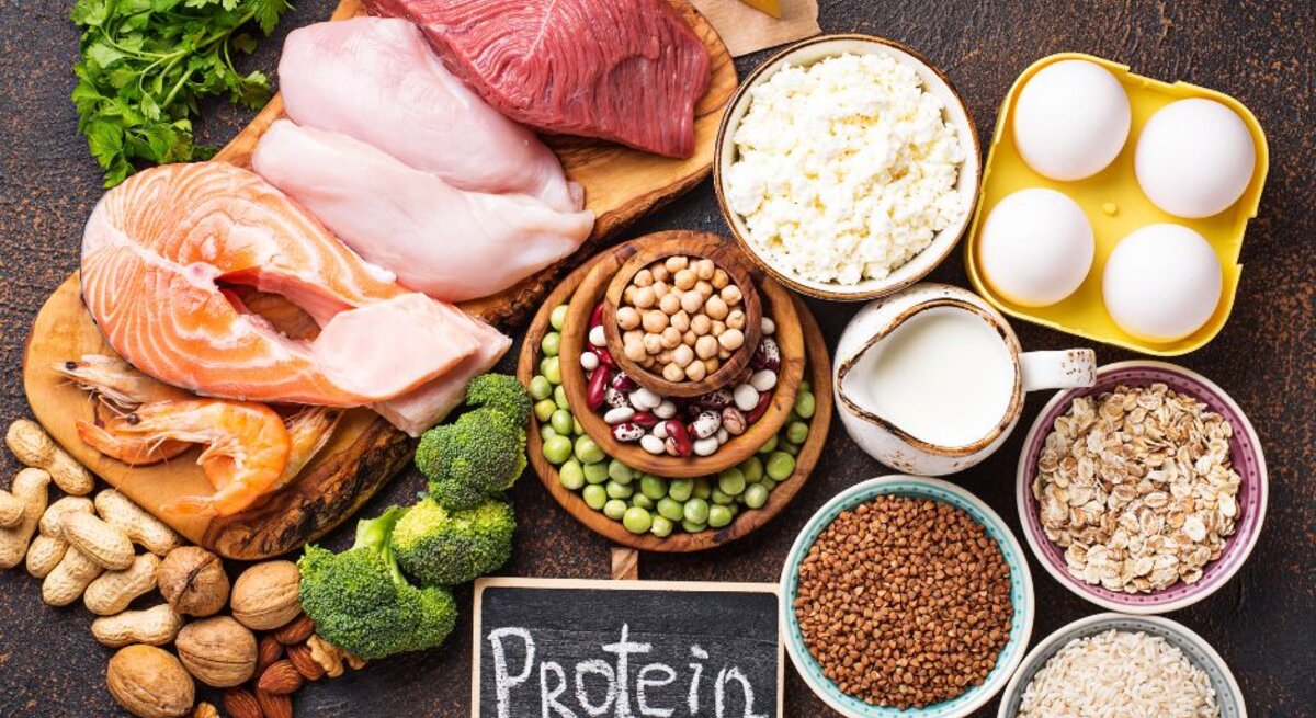 Vielseitiges Arrangement proteinreicher Lebensmittel von tierischer und pflanzlicher Herkunft: rohes Rindfleisch, Hähnchenbrust, Lachssteak und Garnelen, Eier in einer Schale, Käse und Quark, Milch sowie Schalen mit Linsen, Kichererbsen, Bohnen, Erbsen, Haferflocken und Reis; dazu Nüsse, Brokkoli und Kräuter, vor einer kleinen Tafel mit der Aufschrift „Protein“.
